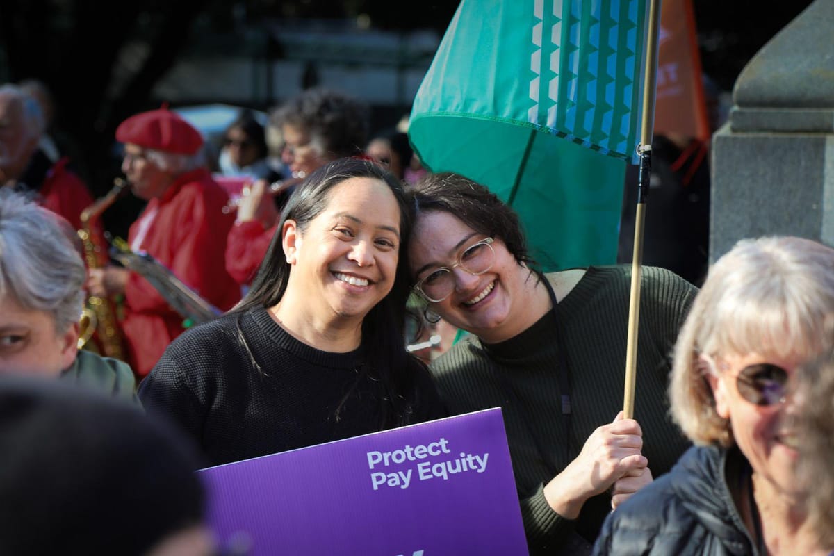 two women smile holding a flag and a purple sign that says "protect pay equity"